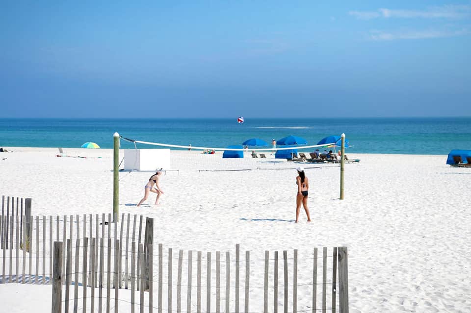 Beach volleyball court with players, ocean view, white sand, and blue umbrellas