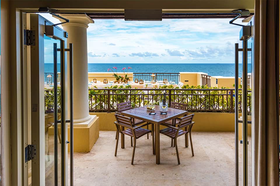 Beachfront balcony with ocean view, dining table, columns, and yellow buildings in distance