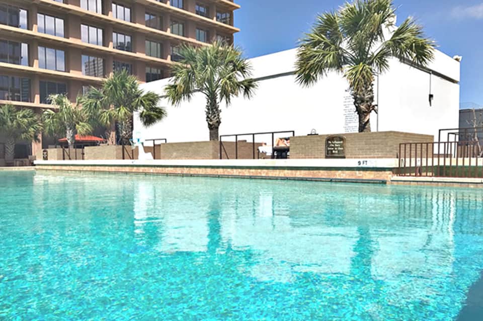 Crystal clear resort pool with palm trees and white building behind sunny blue sky