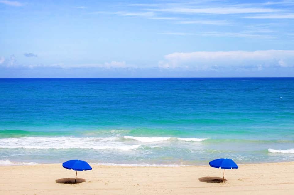 Two blue beach umbrellas on sandy shore with turquoise ocean waves and clear sky