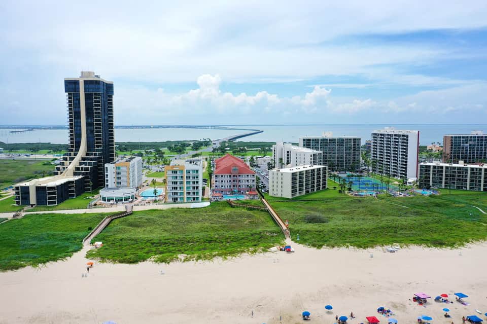 Aerial view of beachfront resort with high-rise hotel tower, multiple buildings, and beach with umbrellas