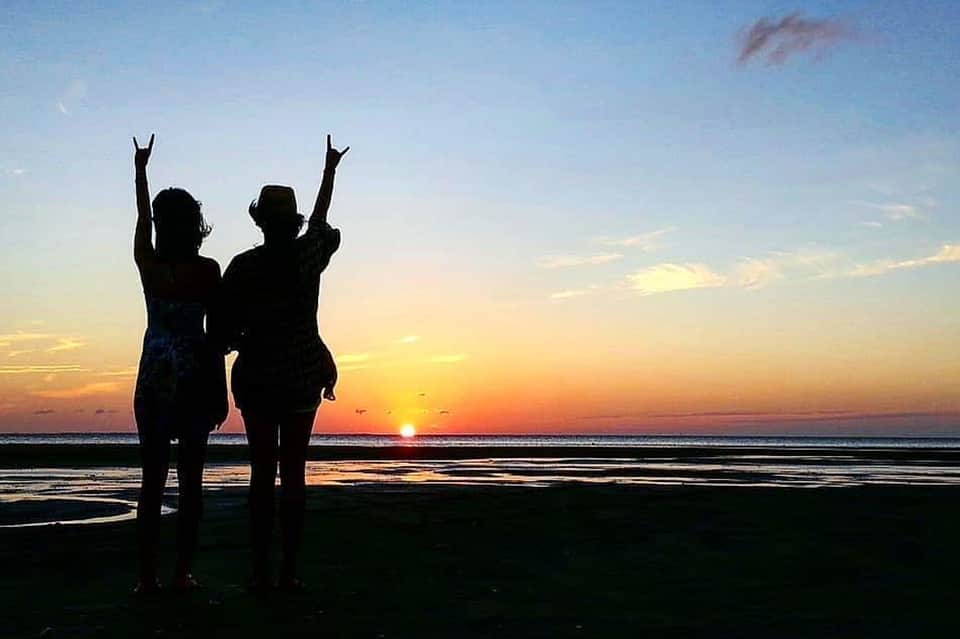 Two people silhouetted against sunset on beach with arms raised in celebration