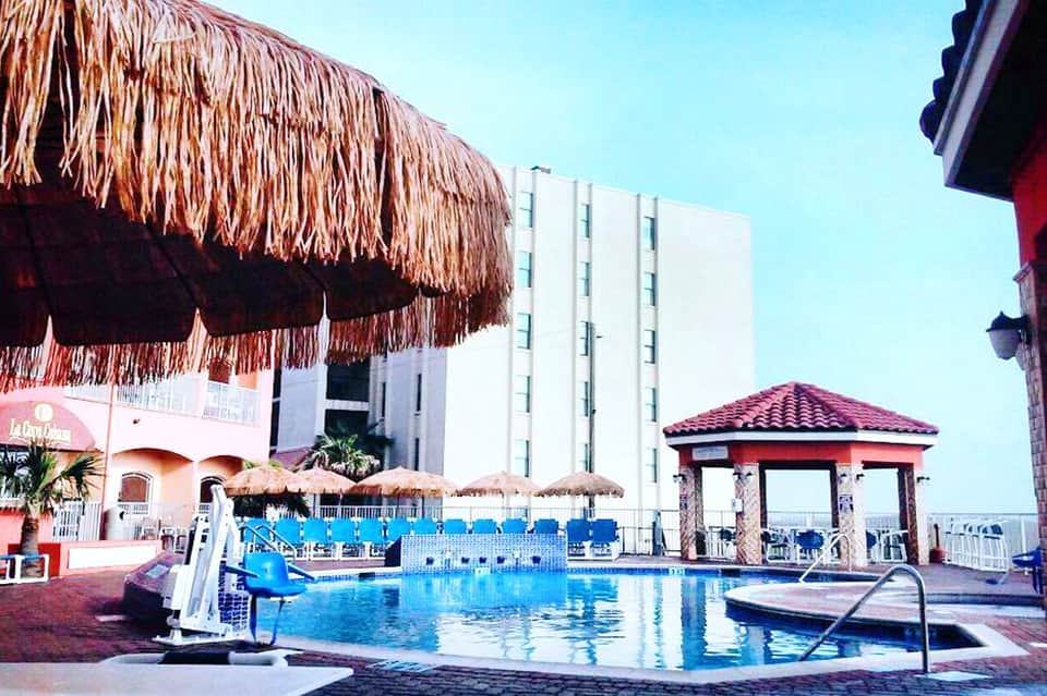 Beachfront resort pool with thatched umbrellas, pink building, and blue lounge chairs