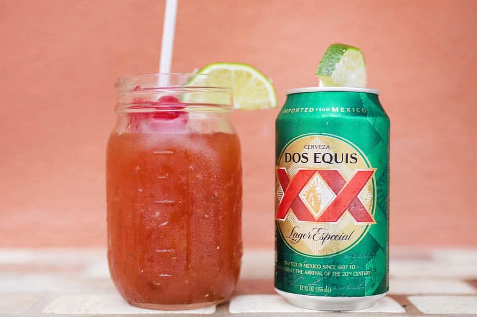 Michelada cocktail with lime garnish next to Dos Equis beer can on coral background