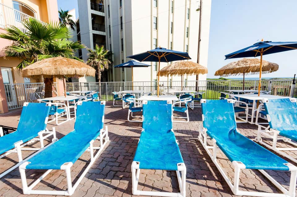 Beach resort pool deck with blue loungers, tiki umbrellas, and colorful beachfront buildings with palm trees