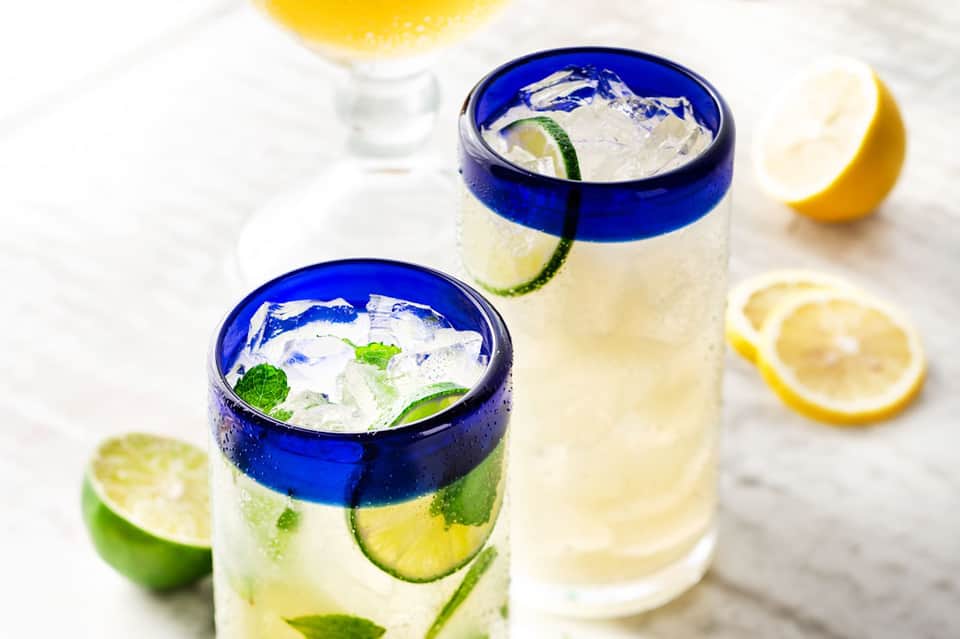 Two refreshing cocktails with lime and lemon slices, ice, and mint garnish in blue-rimmed glasses.