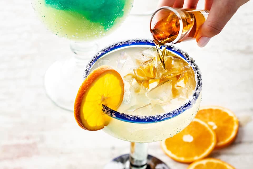 Hand pouring tequila into margarita with blue salt rim and orange slice