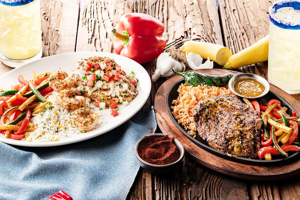 Two plates of grilled seafood with rice and vegetables, alongside fresh lemons and red pepper on rustic wooden table