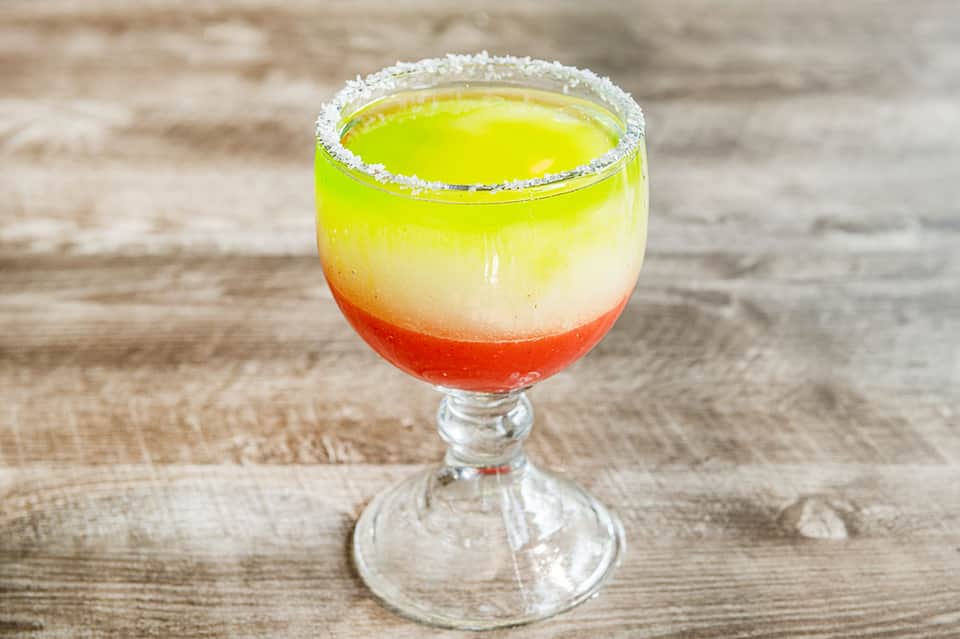 Colorful layered margarita cocktail with salt rim in glass on wooden surface