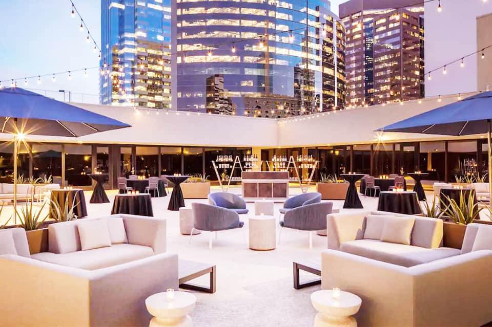 Rooftop evening event space with illuminated city skyline, cream seating areas, and string lights