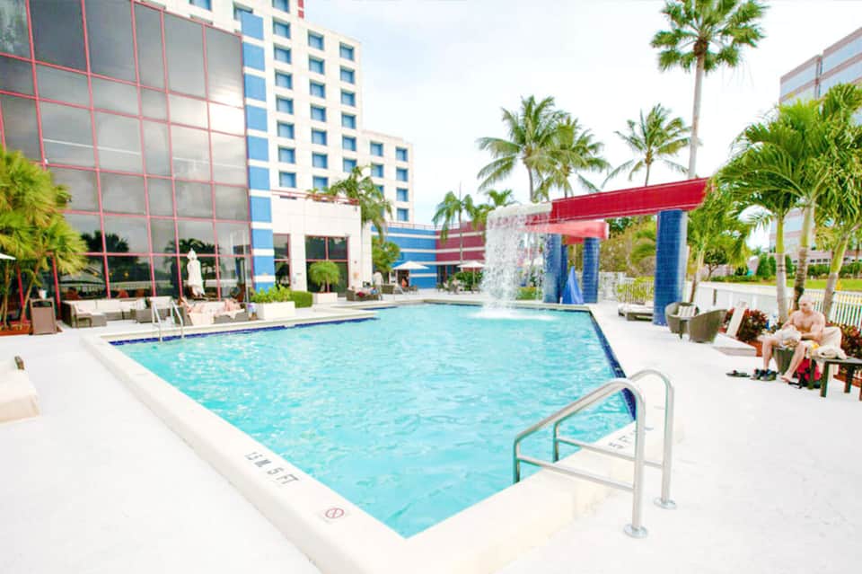 Resort swimming pool with palm trees, red pergola, lounge chairs, and colorful modern buildings