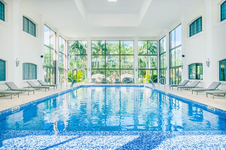 Indoor lap pool with white columns, large windows overlooking palm trees, and gray loungers