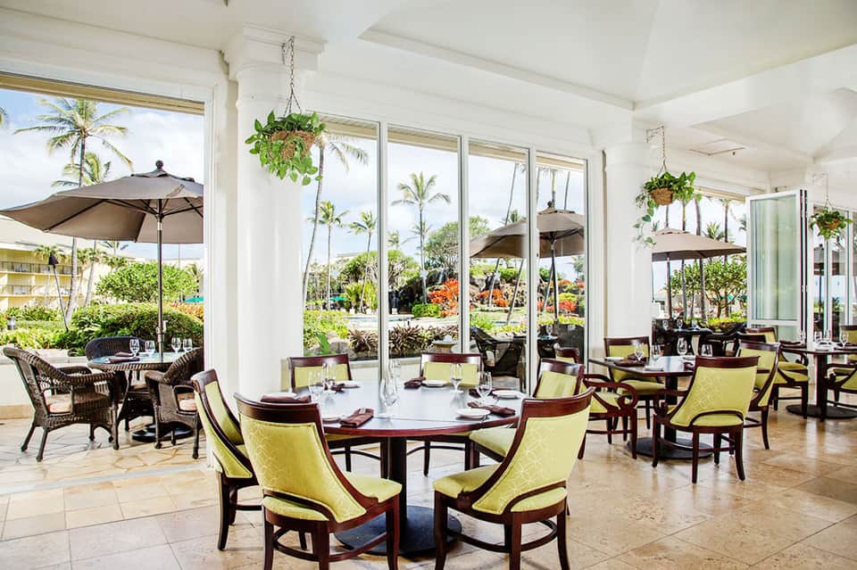 Waterfront dining room with yellow upholstered chairs, palm trees visible through expansive glass doors