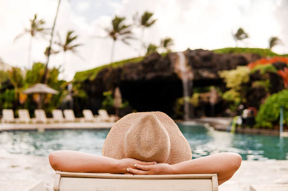 Person wearing straw hat relaxing in lounge chair beside resort pool with palm trees
