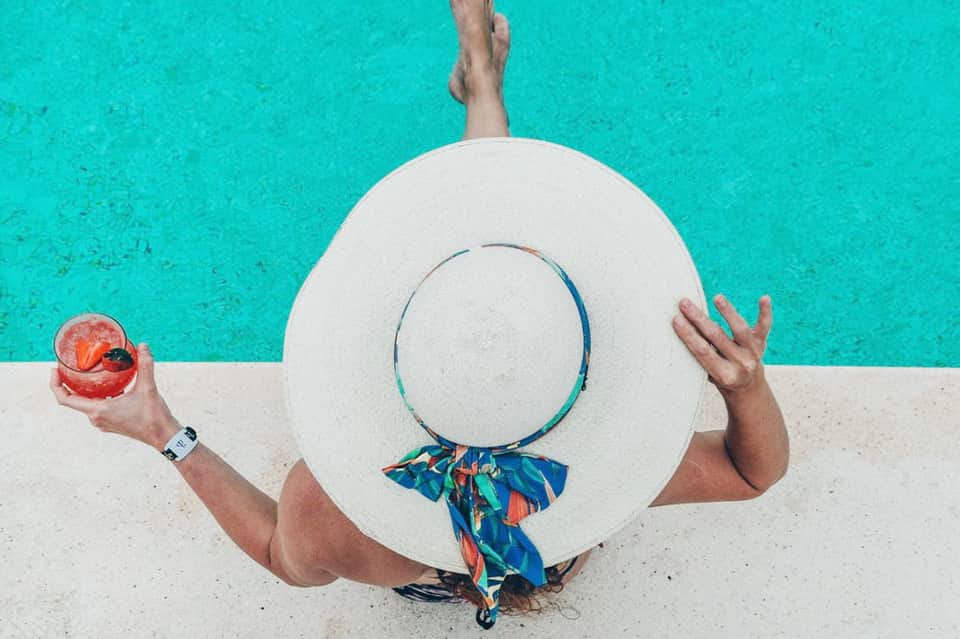 Person in turquoise water holding white sun hat and colorful drink, poolside shot