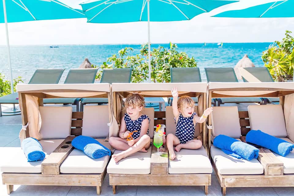 Two young children sitting in wooden cabana beds with ocean view and blue umbrellas at beachfront resort