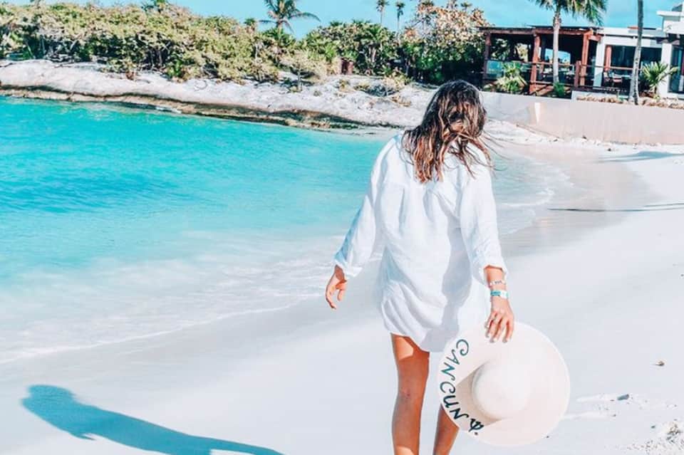 Woman in white shirt walking on white sand beach toward turquoise ocean water