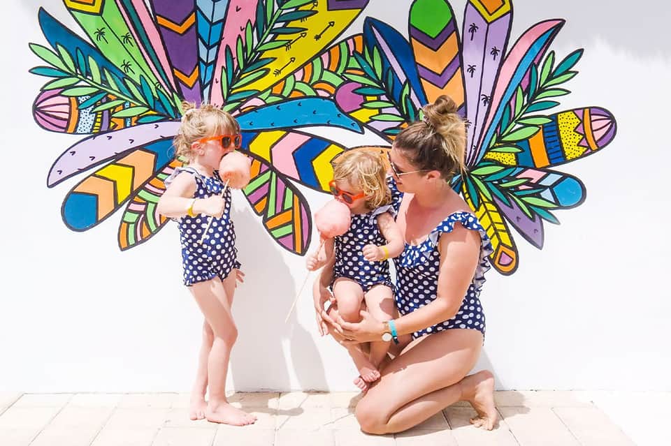Woman and two young children in matching navy swimsuits posing before colorful mural wall art