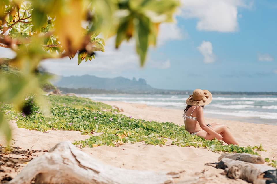 Woman in white bikini sitting on sandy beach under tropical vegetation with ocean and mountains in background