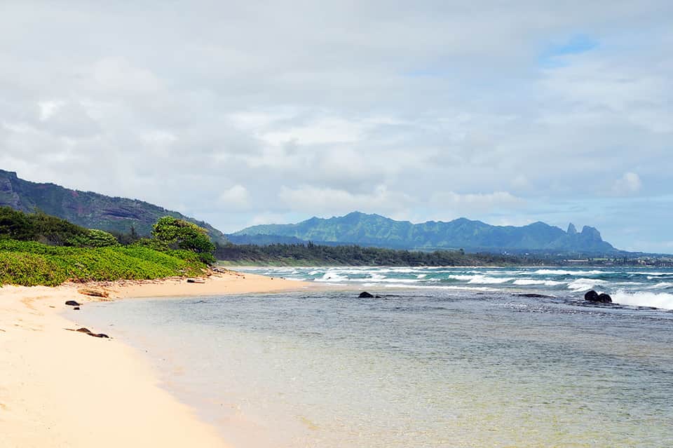 Scenic beach with golden sand, turquoise water, mountains in distance, and lush green vegetation