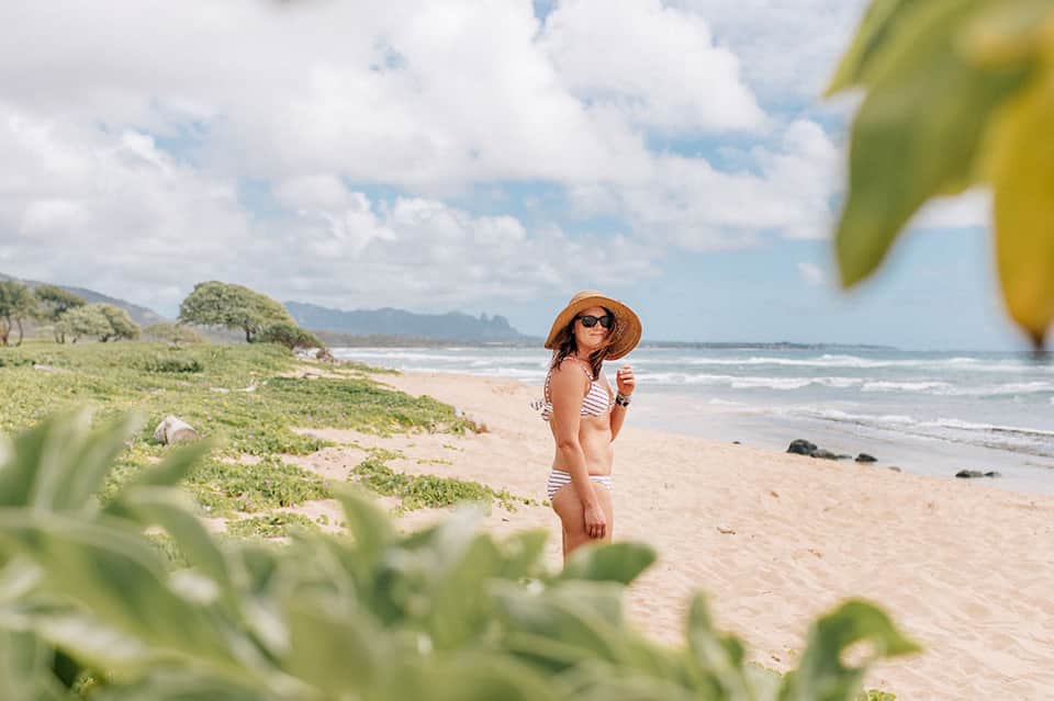 Woman in striped swimsuit standing on sandy beach with tropical vegetation and ocean waves