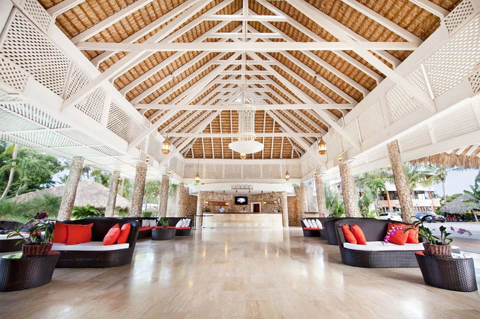 Bright resort lobby with white wooden beams, thatched ceiling, black seating, and red accent pillows