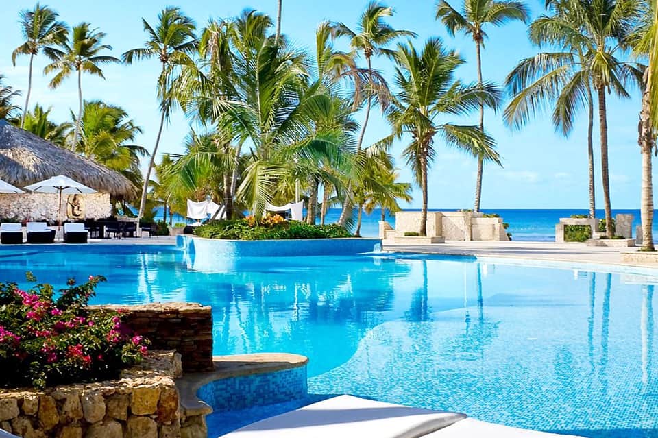 Large resort pool with infinity edge overlooking ocean, palm trees, thatched umbrellas, and clear blue sky