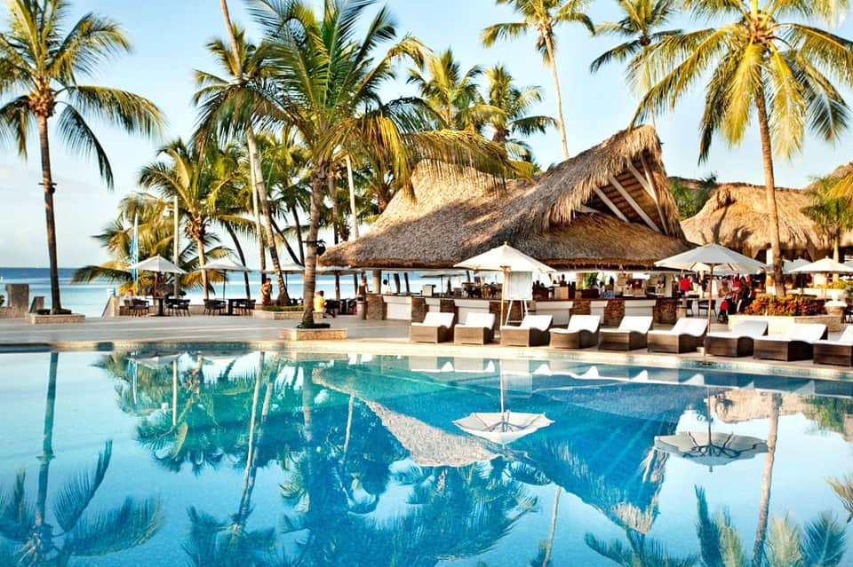 Tropical beachfront resort pool with thatched roof structures, palm trees, and ocean view