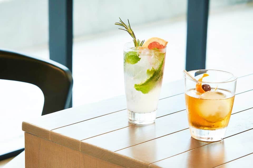 Two cocktails on wooden table by window: mojito with rosemary sprig and grapefruit, and whiskey drink with cherry garnish