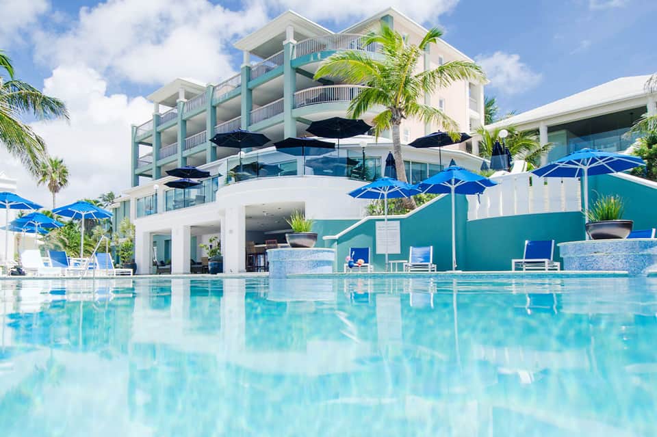 Beachfront resort pool with turquoise water, blue umbrellas, white loungers, and multi-story building with palm trees