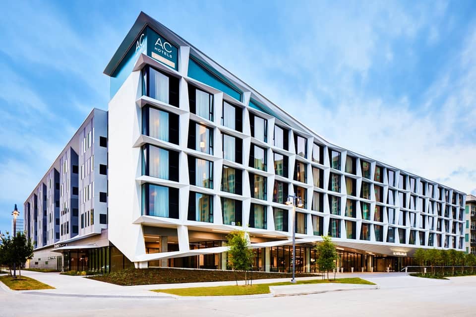 Modern AC Hotel exterior with white facade, grid-pattern windows, and ground-level entrance with landscaping