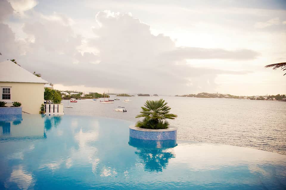 Infinity pool overlooking calm bay with sailboats, palm tree planter in pool, white building and cloudy sky