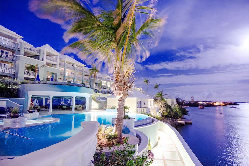 Luxury waterfront resort pool at dusk with palm trees, white building, and blue water reflection