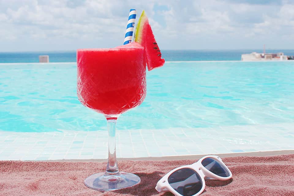 Frozen red cocktail with pineapple garnish on beach near turquoise pool and white sunglasses
