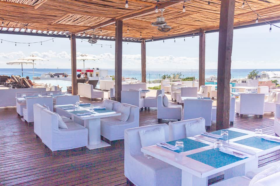 Beachfront lounge area with white furniture, wooden pergola, ocean view, and umbrella