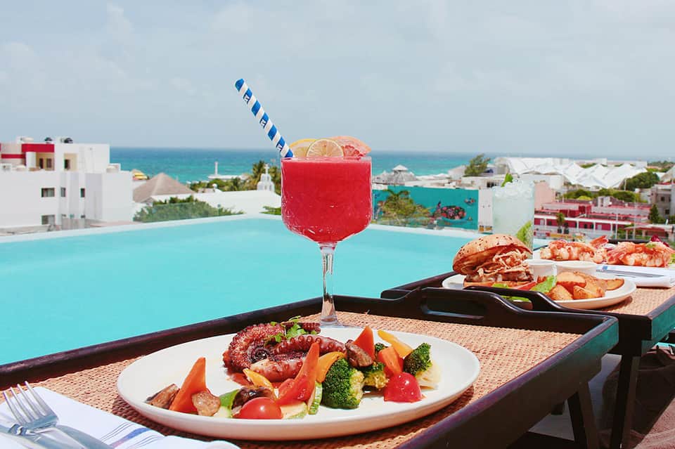 Gourmet plate with drink overlooking infinity pool and ocean, rooftop dining with coastal view