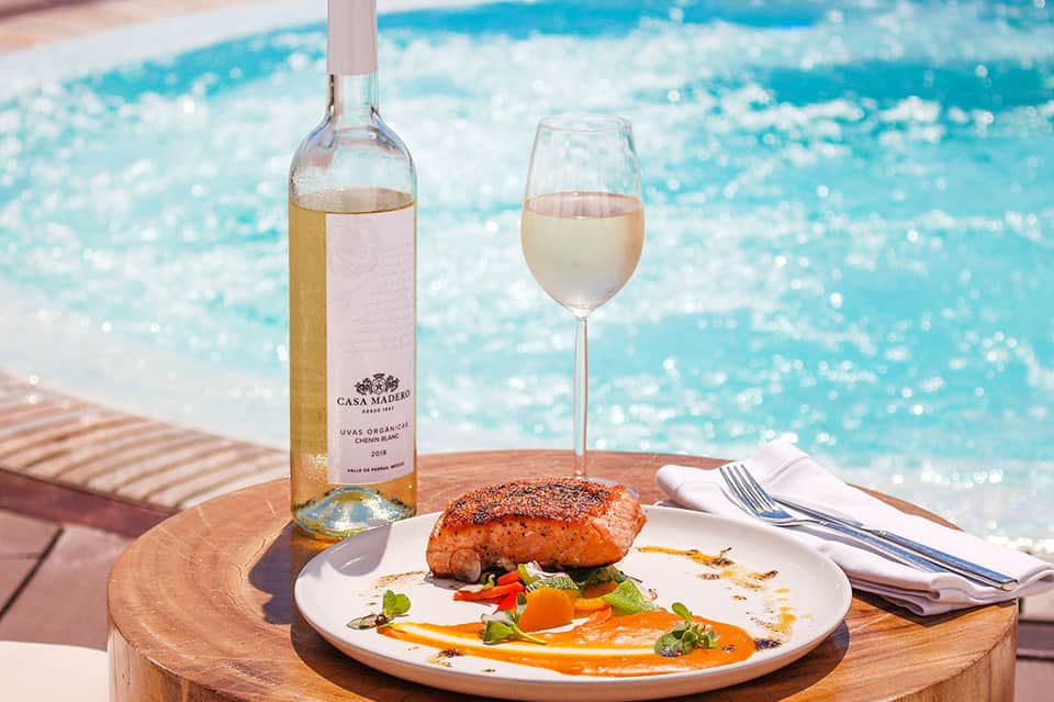 Poolside dining with rosé wine, plated salmon dish, and turquoise water backdrop