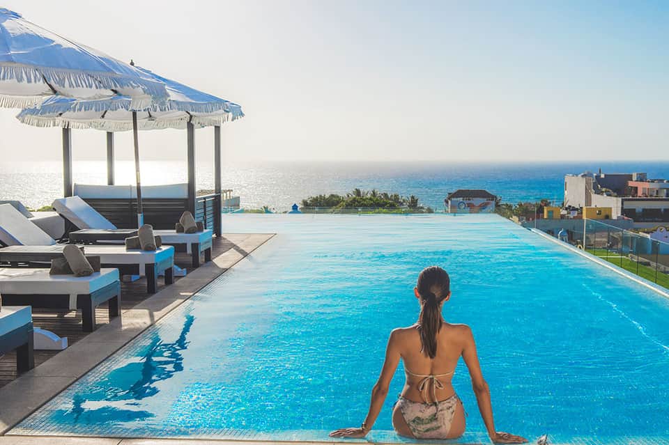 Woman in bikini sitting at edge of infinity pool overlooking tropical ocean coastline