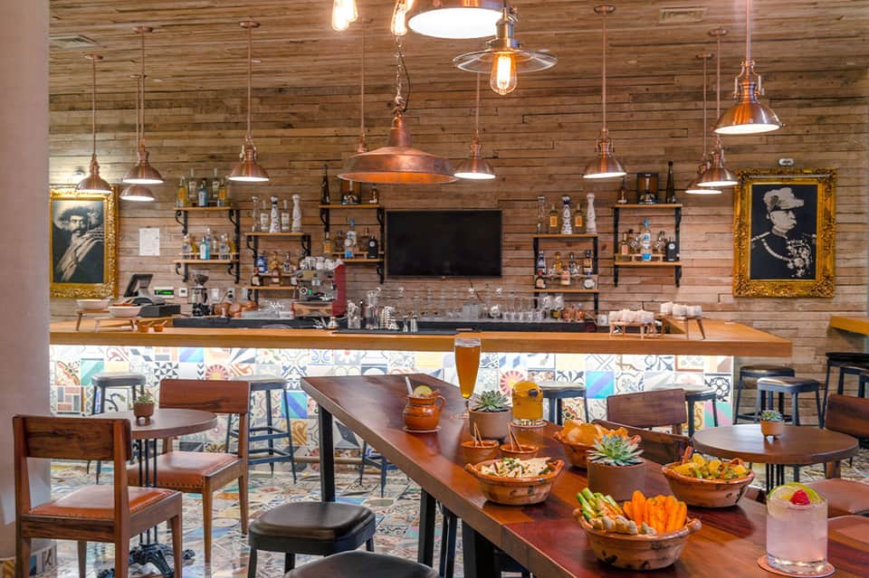 Rustic bar interior with wooden walls, copper pendant lights, and colorful tiled counter