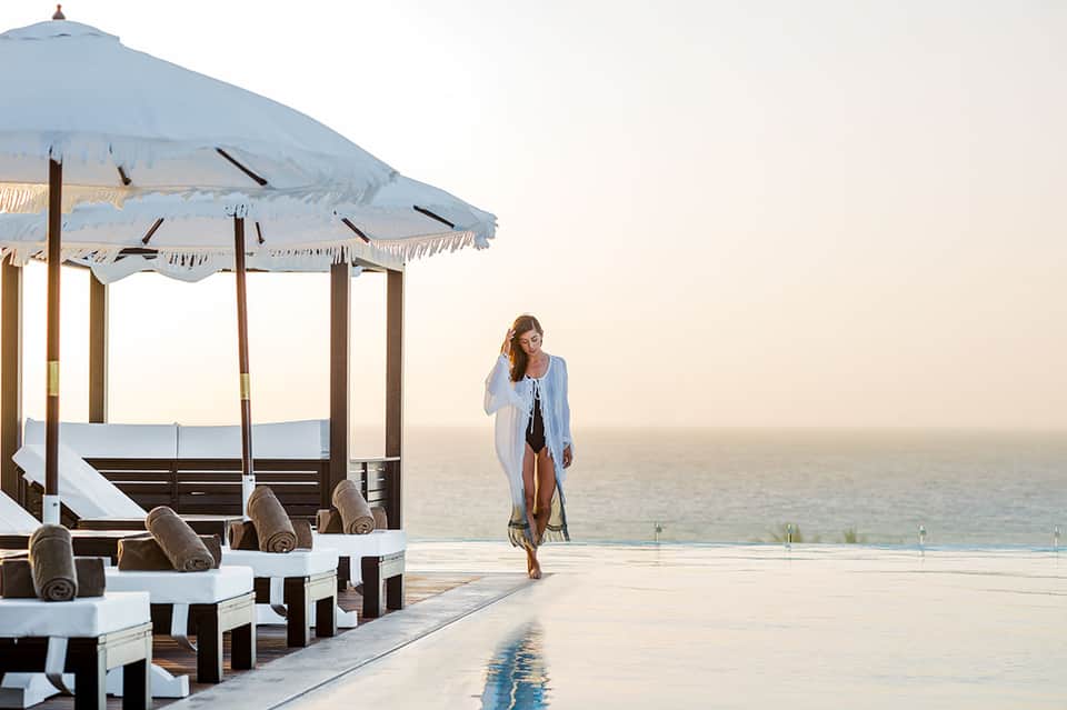 Woman in white cardigan walking by infinity pool overlooking calm ocean at dusk with blue umbrella cabana