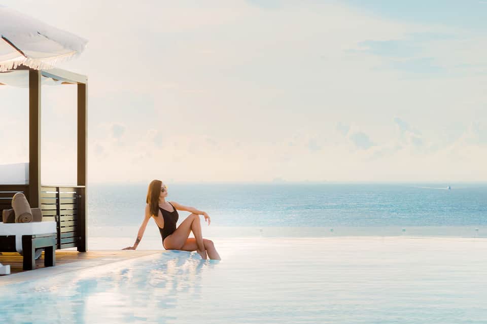 Woman in black swimsuit relaxing by infinity pool overlooking calm turquoise ocean and sky