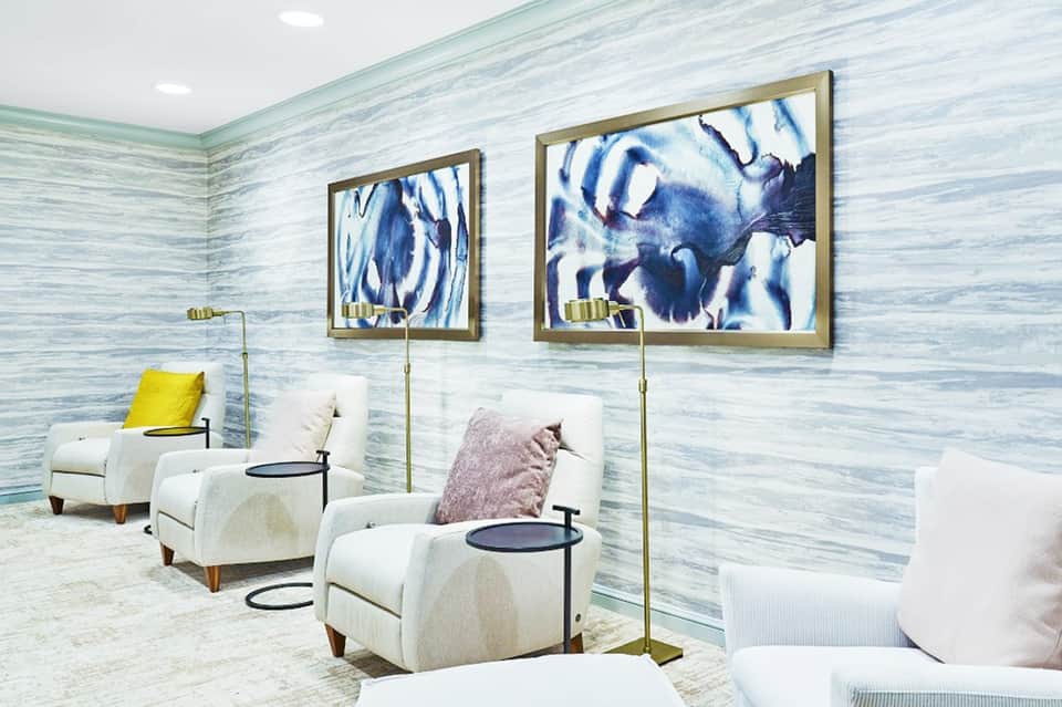 Modern spa lounge with blue abstract artwork, white armchairs, brass floor lamps, and striped wallpaper