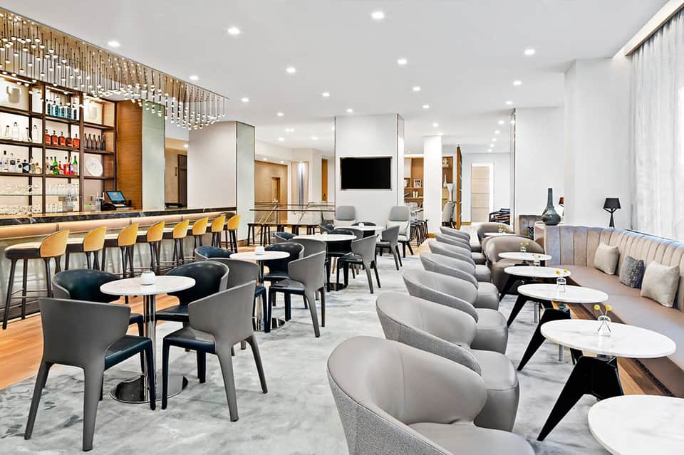 Modern hotel bar and lounge with gray seating, white tables, and wooden accents