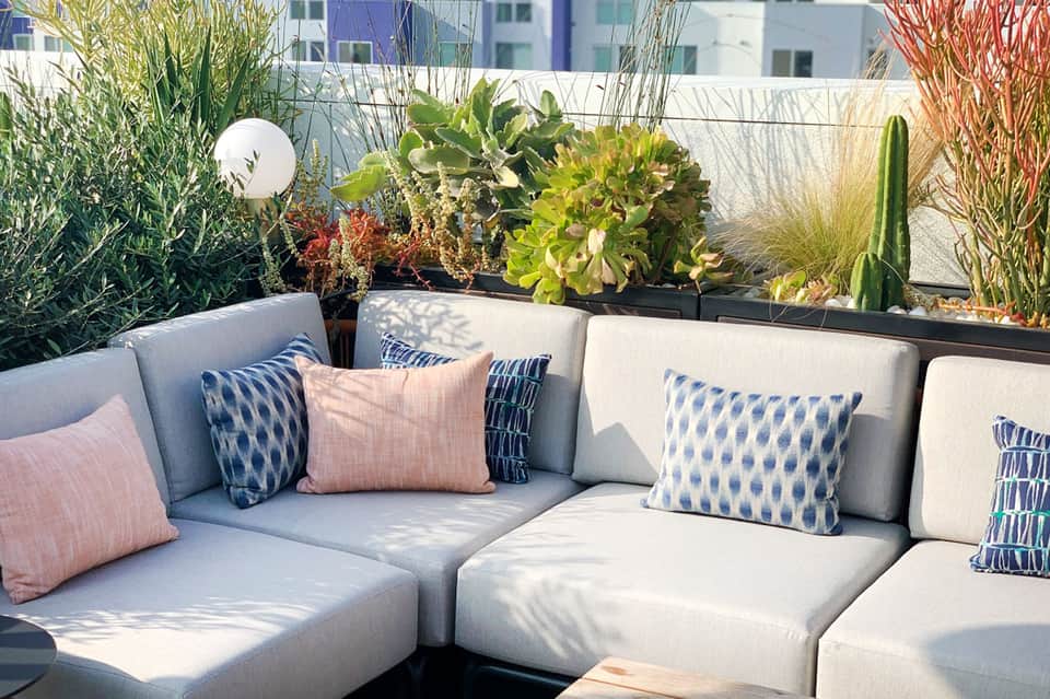 Rooftop seating area with cream cushioned modular sofa, patterned pillows, and lush green plant containers