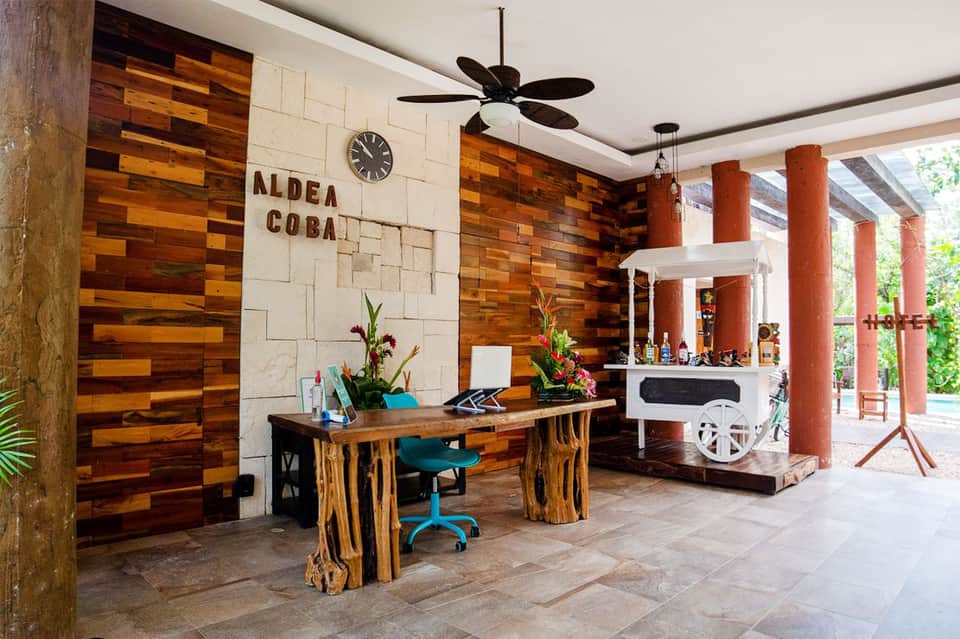 Modern hotel lobby with wooden wall panels, reception desk, red columns, and tropical flowers