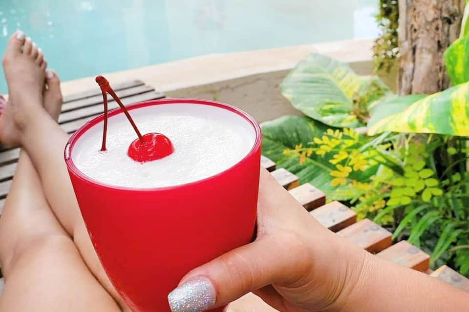 Red cocktail with white creamy top and cherry garnish by poolside with loungers visible