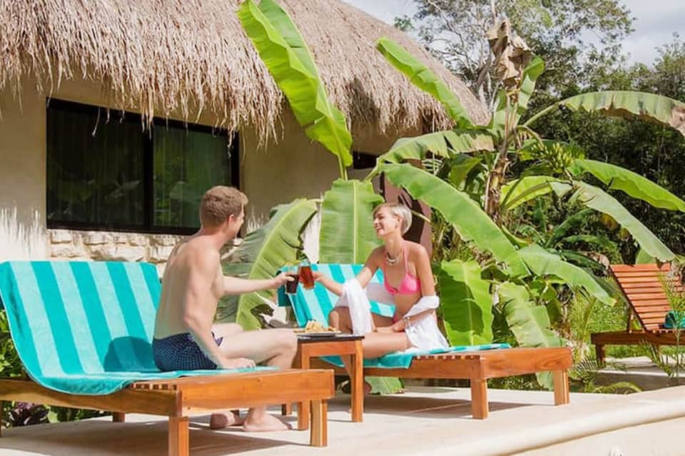 Couple relaxing on turquoise lounge chairs with tropical plants and thatched-roof bungalow in background