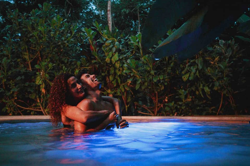 Couple in illuminated indoor pool surrounded by tropical foliage at night