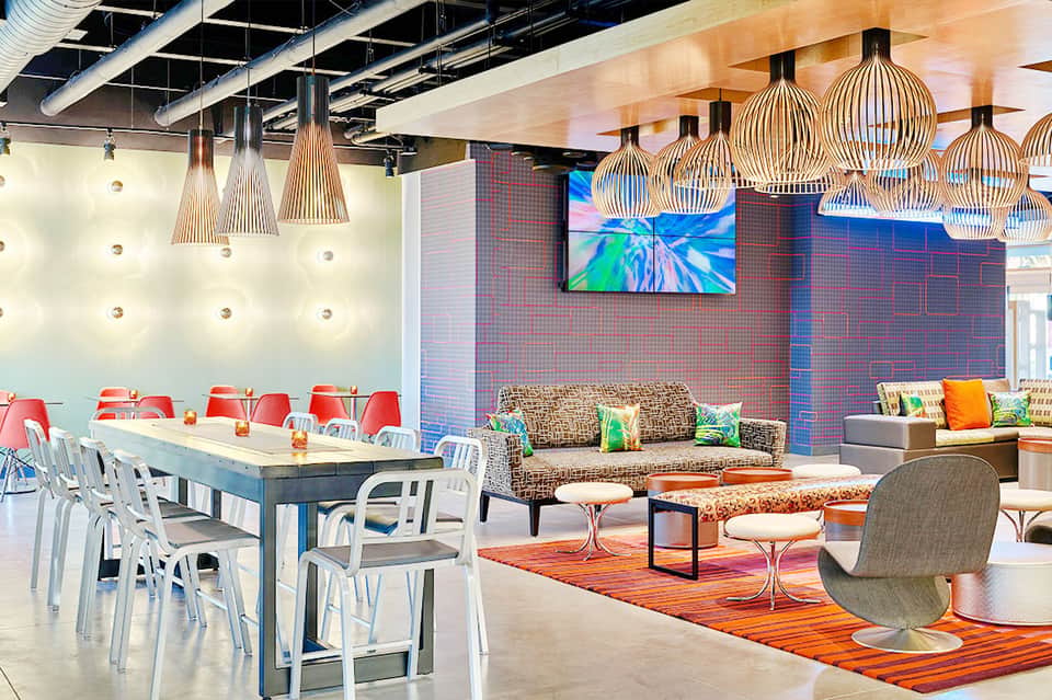 Modern hotel lounge with colorful pendant lights, dining table, patterned sofa, and digital display wall
