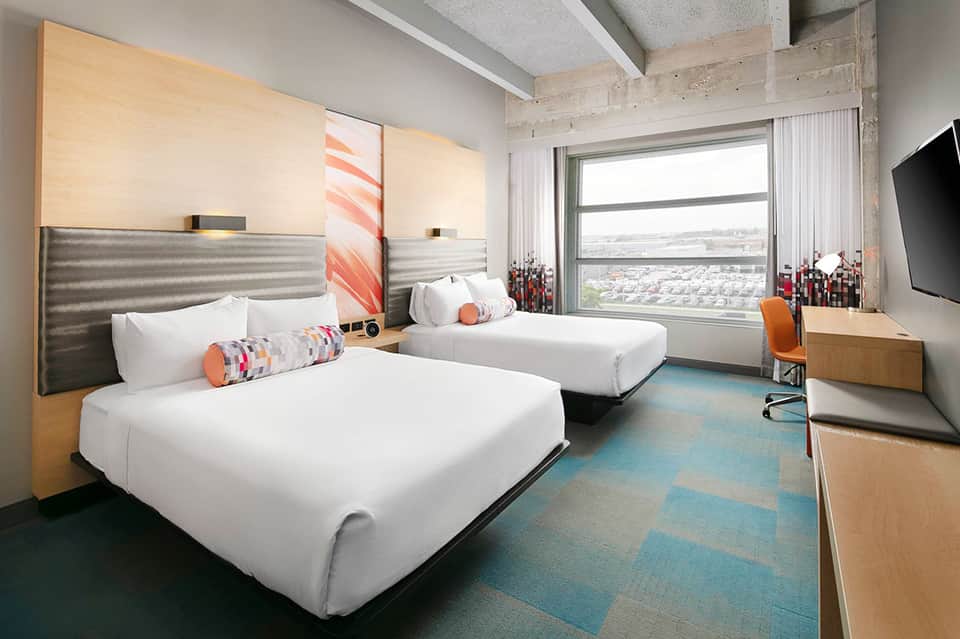 Modern hotel bedroom with two queen beds, wood headboards, and city view windows